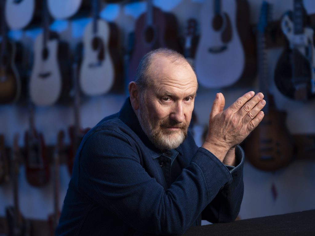 Colin Hay seated, leaning on a counter in front of a background filled with guitars.