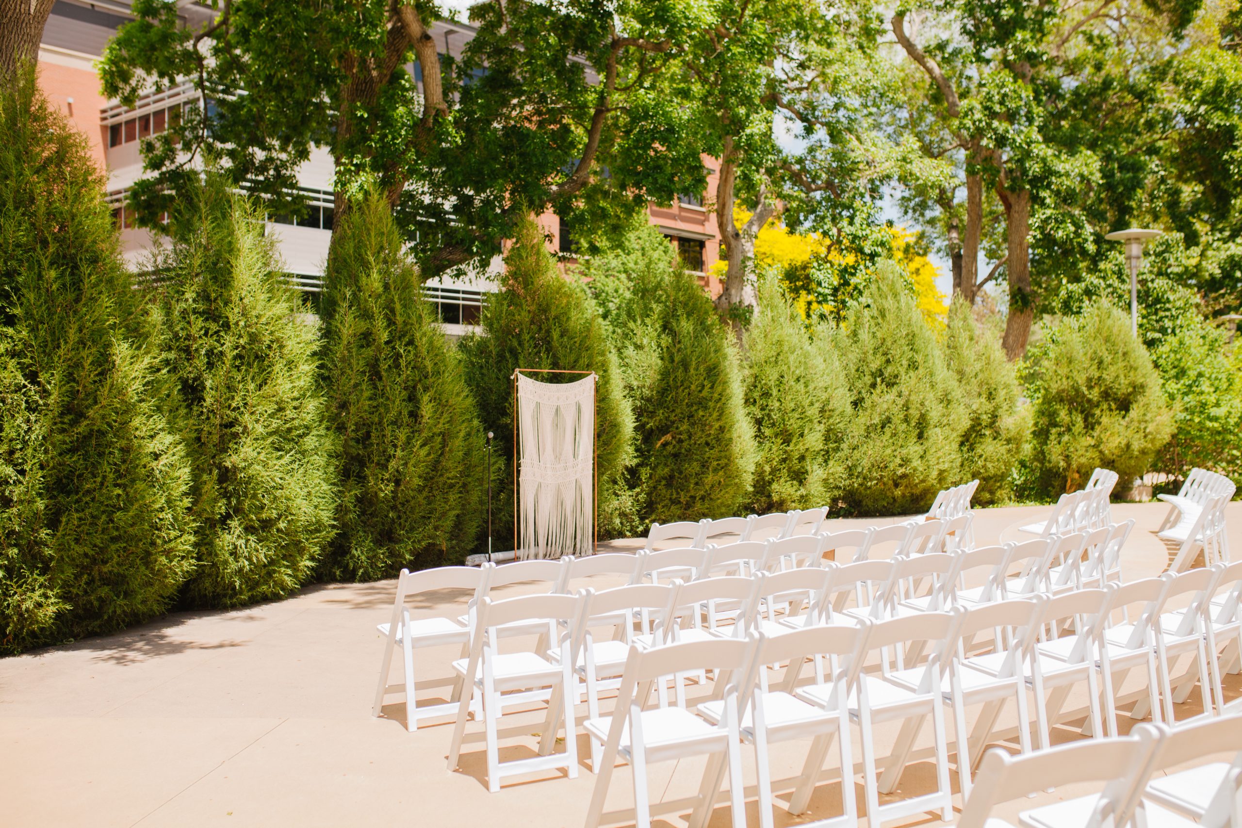 A setup for a wedding on the Terrace with a macrame altar and white charirs. It's sunny and bright, and the trees are tall and green.