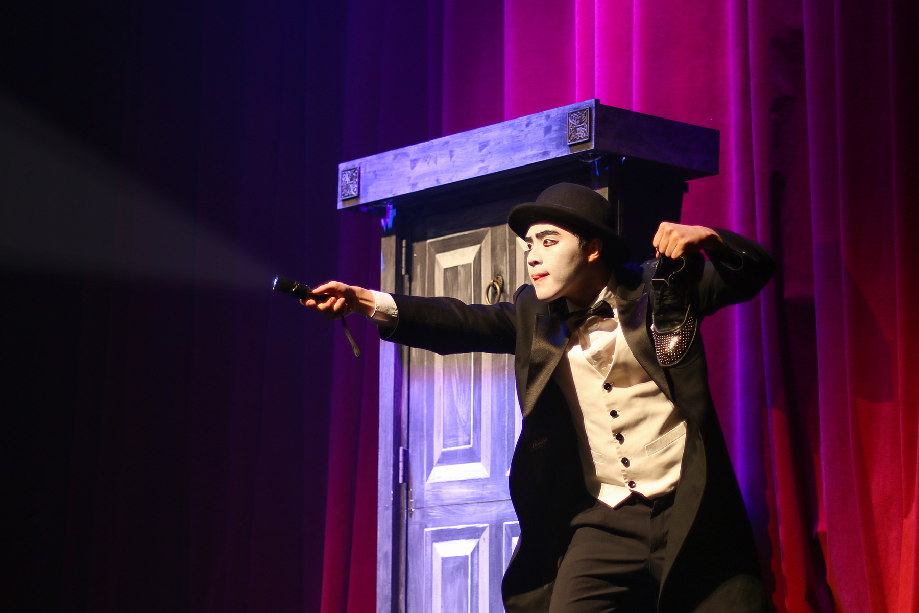 A man wearing white make up dramatically pointing a flashlight next to a door that goes through space and time.