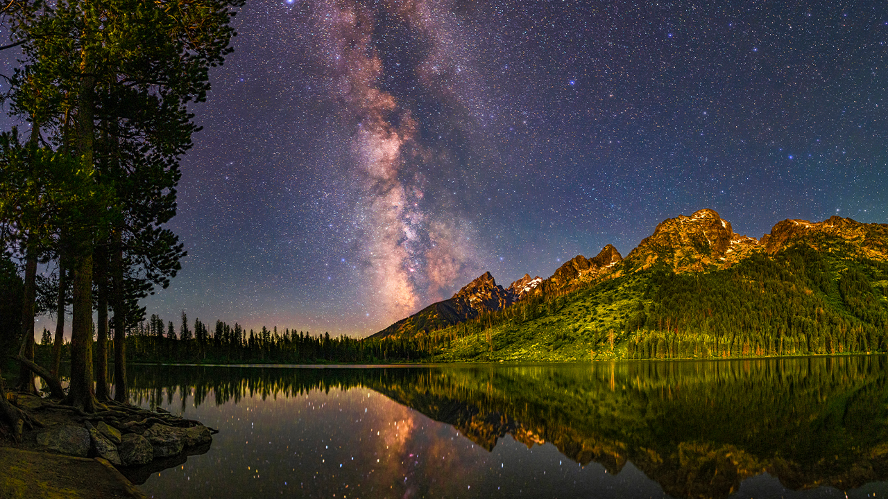 The night sky with a clear view of the Milky Way reflected over a lake and mountains
